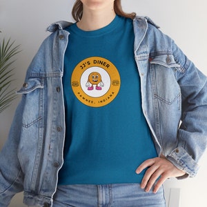 May include: A teal blue t-shirt with a yellow and orange circular graphic featuring a smiling waffle with pink shoes. The graphic says "JJ's Diner" with the text "Famous Waffles" and "Served All Day" on the top and bottom of the circle. The text "Pawnee, Indiana" is below the circle.