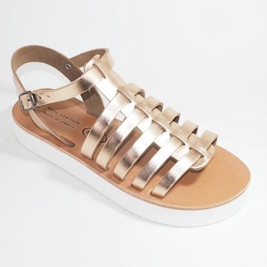May include: Gold-colored leather sandals with a woven strap design. The sandals have a white sole and a buckle closure. The sandals are labeled "REAL LEATHER MADE IN GREECE".