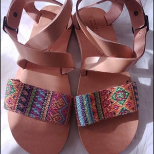 Könnte beinhalten: Ein Paar hellbraune Ledersandalen mit farbenfrohen gewebten Riemen. Die Sandalen haben Knöchelriemen mit schwarzen Schnallen. Die gewebten Riemen weisen geometrische Muster in Rosa-, Blau-, Orange- und Grüntönen auf.