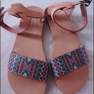 Könnte beinhalten: Ein Paar hellbraune Ledersandalen mit bunten gewebten Stoffriemen. Die Sandalen haben ein schlichtes Design mit Knöchelriemen und Schnallen. Die Fußbetten sind mit Text bedruckt. Die Sandalen werden auf einer weißen Oberfläche präsentiert.
