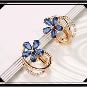 Könnte beinhalten: Zwei goldfarbene Ohrringe in Blumenform. Jeder Ohrring hat eine blaue Edelsteinblume mit einem kleinen weißen Edelstein in der Mitte. Die Ohrringe haben ein mehrschichtiges Design mit einer Reihe kleiner weißer Edelsteine.