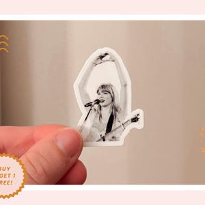 K&ouml;nnte beinhalten: Ein schwarz-wei&szlig;er Aufkleber mit einer S&auml;ngerin mit Mikrofon und Gitarre. Der Aufkleber hat einen wei&szlig;en Rand und wird von einer Hand gehalten. Der Aufkleber hat den Text "BUY 3, GET 1 FREE!" auf einem goldenen Aufkleber.
