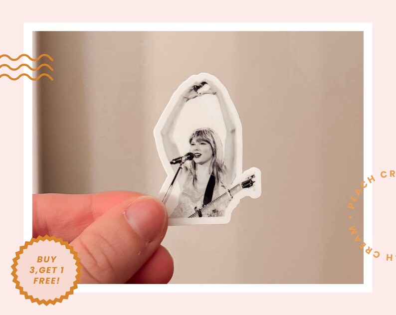 K&ouml;nnte beinhalten: Ein schwarz-wei&szlig;er Aufkleber mit einer S&auml;ngerin mit Mikrofon und Gitarre. Der Aufkleber hat einen wei&szlig;en Rand und wird von einer Hand gehalten. Der Aufkleber hat den Text "BUY 3, GET 1 FREE!" auf einem goldenen Aufkleber.