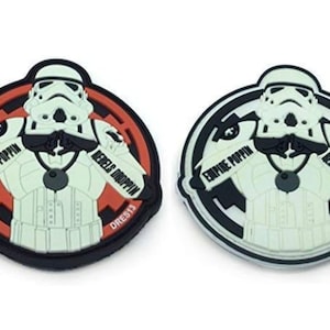 Può includere: Due toppe rotonde nere e bianche con una figura di Stormtrooper bianca al centro. Lo Stormtrooper indossa una catena d'oro e ha il testo "Empire Poppin'" su un braccio e "Rebels Droppin'" sull'altro. Il testo "DREST13" è in basso su ogni toppa.