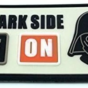 Peut inclure: Écusson rectangulaire avec l'inscription "THE DARK SIDE", des boutons "OFF" et "ON", et un dessin du casque de Dark Vador. Le patch a une bordure noire, un fond beige clair et est destiné aux vêtements ou accessoires.
