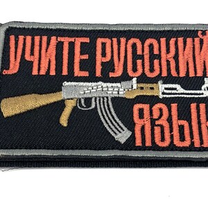 May include: A rectangular embroidered patch with a black background and a grey border. It features the orange Cyrillic text "УЧИТЕ РУССКИЙ ЯЗЫК" and a central graphic of a rifle with a tan stock and a silver-coloured barrel and magazine.
