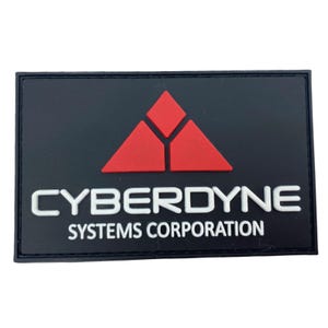 Op de afbeelding: Zwarte patch met een rood driehoekig logo en de tekst "CYBERDYNE SYSTEMS CORPORATION" in wit.