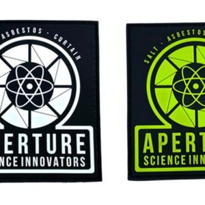 Op de afbeelding: Twee zwarte stoffen patches met het Aperture Science-logo. Het logo toont een gestileerd atoomsymbool in een cirkel met de tekst "SALT - ASBESTOS - CURTAIN" rond de cirkel. De tekst "APERTURE SCIENCE INNOVATORS" staat onder de cirkel. Eén patch heeft een wit atoomsymbool en een witte cirkel, de andere heeft een groen atoomsymbool en een groene cirkel.