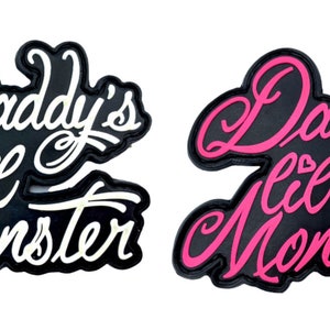 Peut inclure: Deux patchs thermocollants noirs avec des lettres blanches et roses. Les patchs indiquent "Daddy's Lil Monster".