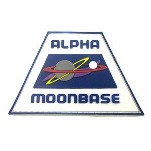 Peut inclure: Un écusson triangulaire bleu et blanc avec le texte "ALPHA MOONBASE" et une image stylisée d'une planète avec des anneaux.