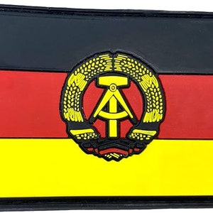 May include: A black, red and yellow patch with a black border. The patch features a stylised hammer and compass inside a wreath of wheat. The patch is a representation of the flag of the German Democratic Republic.