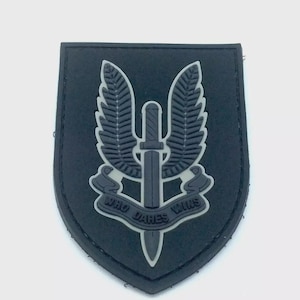 May include: A black and grey patch in a shield shape, featuring a central sword design with wings on either side. The text "WHO DARES WINS" is displayed on a banner below. This patch is likely an emblem or insignia.