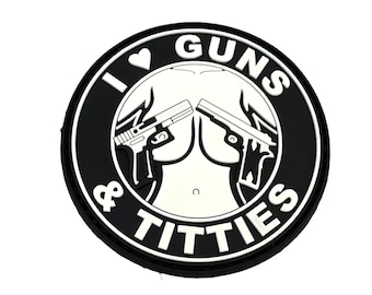 May include: A circular black and white patch featuring a graphic illustration of two handguns positioned over a stylized torso. The text "I HEART GUNS & TITTIES" is printed in bold white capital letters around the border of the circular design.