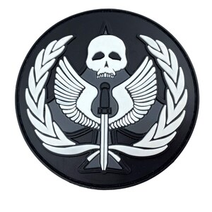 May include: A circular black and white morale patch featuring a central white skull positioned above a vertical dagger with outstretched wings. The entire design is framed by a white laurel wreath set against a solid black circular background.