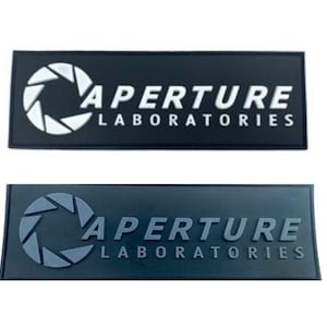Op de afbeelding: Twee zwarte rechthoekige patches met het Aperture Laboratories-logo. Het logo is een witte cirkel met een gestileerde camera lens erin en de tekst "Aperture Laboratories" in witte letters. De bovenste patch heeft een wit logo en de onderste patch heeft een grijs logo.