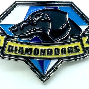 May include: A silver metal pin featuring a black diamond shape. Inside the diamond is a black silhouette of a dog's head. Below the diamond is a yellow banner with the text "DIAMOND DOGS".