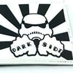 Peut inclure: Un écusson rectangulaire blanc avec un motif noir et blanc. Il présente un casque de Stormtrooper avec des lunettes de soleil, sur fond de rayons noirs et blancs. Les mots "DARK SIDE" sont tatoués sur les mains.