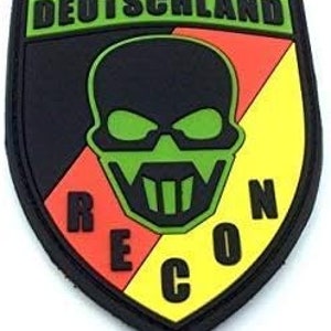 May include: A black, yellow, red and green embroidered patch with a skull and crossbones design. The skull is green with a black mask. The text "RECON" is written in black letters on the patch. The text "DEUTSCHLAND" is written in green letters at the top of the patch.