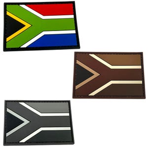 Może przedstawiać: Trzy naszywki z flagą Republiki Południowej Afryki w różnych schematach kolorystycznych. Pierwsza naszywka jest w tradycyjnych kolorach: czerwonym, żółtym, zielonym, niebieskim i czarnym. Druga naszywka jest w kolorze brązowym, beżowym i czarnym. Trzecia naszywka jest w kolorze szarym, białym i czarnym.