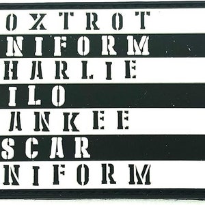 May include: Black and white patch with the NATO phonetic alphabet spelled out in all capital letters. The letters are arranged in rows, with each row representing a different letter. The letters are white on a black background.