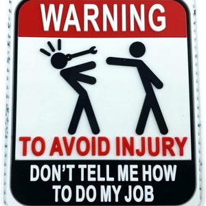 May include: A rectangular patch with a white background, red top section, and black bottom section. The text "WARNING" is in white block letters above stick figures. The text "TO AVOID INJURY" and "DON'T TELL ME HOW TO DO MY JOB" are also in white.