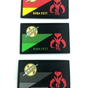 May include: Three black and white patches with a red, green, and grey stripe across the top. The patches have a Mandalorian symbol and the text "BOBA FETT" on them.