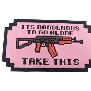 May include: A rectangular patch with a black border and a light pink background. The centre features a pixelated graphic of a rifle in dark grey and orange. The text "ITS DANGEROUS TO GO ALONE" appears above the rifle, and "TAKE THIS" is below.