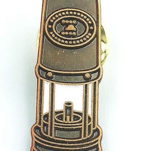 May include: A bronze-coloured metal pin shaped like a miner's lamp. The lamp has a round, cylindrical top with a circular design and a glass-like window. The bottom of the lamp has a round base with a decorative design.