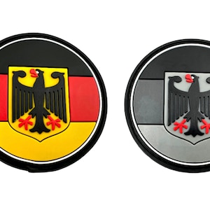 May include: Two round, black-bordered patches featuring a German flag and eagle design. One patch has a yellow, red, and black flag, while the other has a grey, white, and black flag.