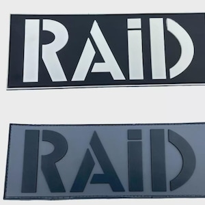 Può includere: Due toppe rettangolari con la scritta "RAID" in grassetto e in stile. La toppa superiore presenta lettere bianche su sfondo nero, mentre la toppa inferiore presenta lettere grigio scuro su sfondo grigio chiaro.
