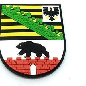 May include: A black and green embroidered patch featuring a black bear walking on a red brick wall with a white archway. The top half of the patch shows a black eagle with yellow beak and talons on a yellow background with black and green stripes. The eagle is surrounded by a green crown.