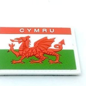 Può includere: Una toppa bianca con un drago rosso e la parola "CYMRU" in lettere rosse. Il drago è su uno sfondo verde e bianco.
