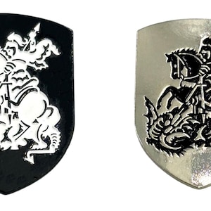 Könnte beinhalten: Zwei metallene Schild-förmige Pins mit einem Schwarzweißbild eines Ritters zu Pferd, der einen Drachen tötet. Der Ritter trägt eine Rüstung und hält ein Schwert. Der Drache ist unter dem Pferd zusammengerollt.