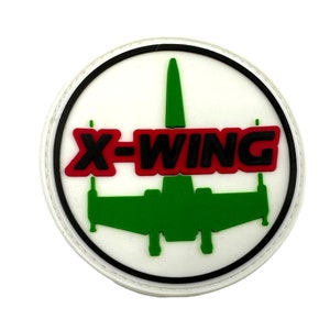 Könnte beinhalten: Ein weißer, runder Patch mit schwarzem Rand. In der Mitte des Patches befindet sich ein grünes X-Wing-Raumschiff mit dem Text "X-WING" in roten und schwarzen Buchstaben.