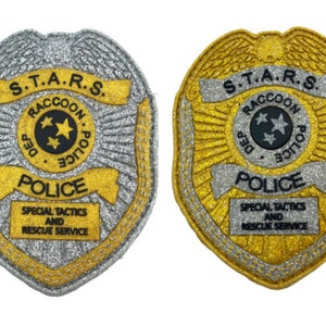 Op de afbeelding: Twee zilveren en gouden politiebadges met de tekst "S.T.A.R.S. Raccoon Police Dept. Police" en "Special Tactics and Rescue Service".