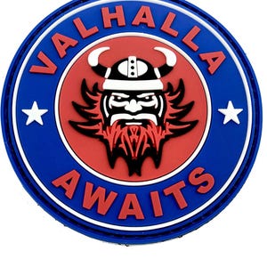 May include: A circular rubber patch featuring a red centre with a black and white Viking head graphic. The blue border contains the white text VALHALLA AWAITS in red lettering, with two white stars on the left and right sides of the design.