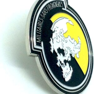 May include: A silver metal pin with a black and yellow design. The pin features a white stylized map of the world with the text "MILITAIRES SANS FRONTIERES" around the outside.