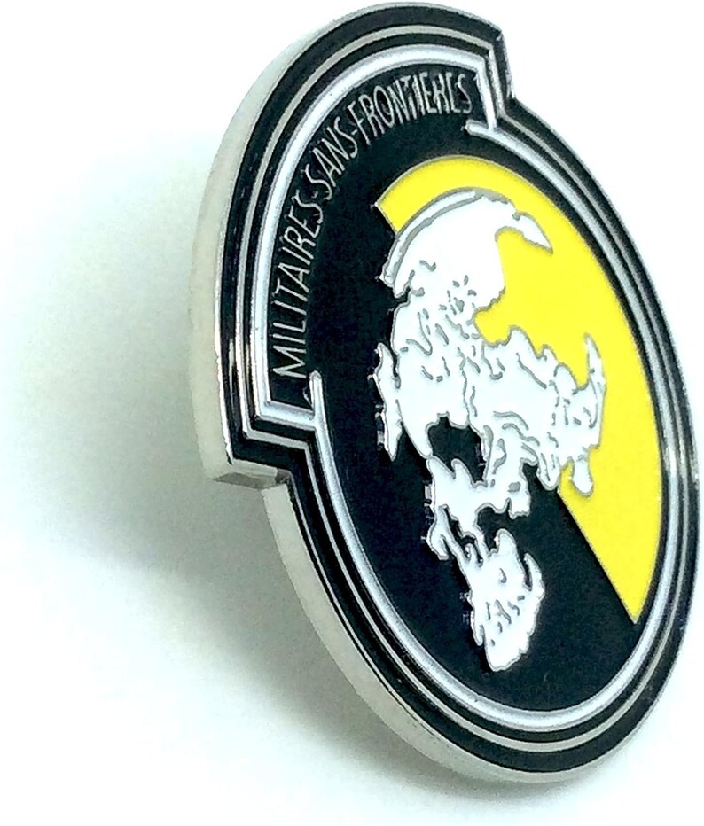 May include: A silver metal pin with a black and yellow design. The pin features a white stylized map of the world with the text "MILITAIRES SANS FRONTIERES" around the outside.