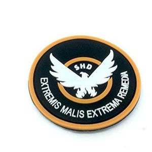 May include: A circular patch featuring a black background with a tan border. In the centre, a white eagle graphic sits above the text SHD. The text EXTREMIS MALIS EXTREMA REMEDIA is printed in white along the bottom curve of the black circular field.