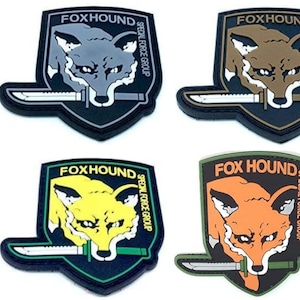 Könnte beinhalten: Vier verschiedene farbige Patches mit einem Fuchs-Kopf und einem Messer. Die Patches sind schwarz mit grünen, gelben, orangefarbenen und grauen Fuchs-Köpfen. Der Text auf den Patches lautet "FOXHOUND" und "SPECIAL FORCE GROUP".