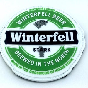 Può includere: Un cerotto circolare bianco, verde e nero con il testo "Winterfell Beer" e "Winter is Coming" in un cerchio attorno a un emblema di lupo grigio. Il testo "Winterfell" è in un grande stendardo nero con lettere bianche. Il testo "Stark" è in uno stendardo più piccolo sotto il grande stendardo. Il testo "Brewed in the North" è in uno stendardo verde sotto lo stendardo Stark. Il testo "Made in the Godswoood of House Stark" è in un cerchio più piccolo attorno all'esterno del cerotto.