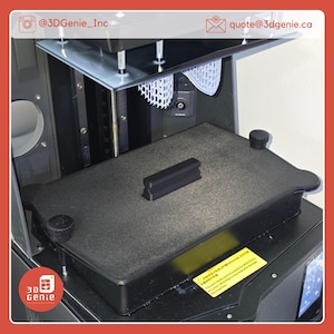 May include: A 3D printer with a black base and a clear top. A black rectangular object sits on the base. The printer has a screen and a white, web-like structure inside. The text "3D Genie" is visible.