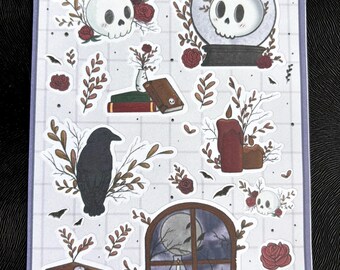 May include: A sheet of stickers featuring a spooky, gothic theme. The stickers include a skull in a snow globe, a raven on a branch, a candle, a window with a full moon, and a stack of books. The stickers are on a white background with a grid pattern.