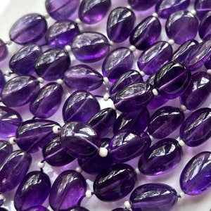 May include: A strand of smooth, oval-shaped amethyst beads in a deep purple hue. The beads are strung on white thread and are perfect for jewelry making.