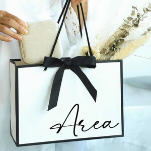 May include: A white gift bag with black trim and handles, tied with a black bow. The word "Area" is written in a cursive font on the front of the bag. The bag contains dried flowers and a small, beige, square box.