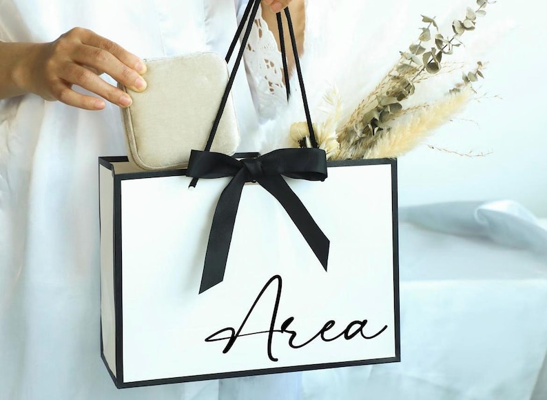 May include: A white gift bag with black trim and handles, tied with a black bow. The word "Area" is written in a cursive font on the front of the bag. The bag contains dried flowers and a small, beige, square box.