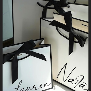 May include: White gift bags with black trim and black satin bows. The bags have handwritten names in black script. The bags are a classic and elegant design, perfect for gifts or special occasions.