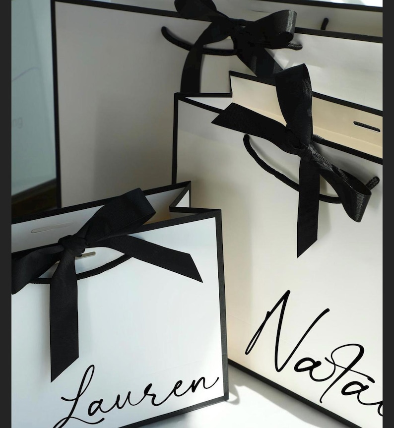 May include: White gift bags with black trim and black satin bows. The bags have handwritten names in black script. The bags are a classic and elegant design, perfect for gifts or special occasions.