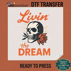 May include: A DTF transfer sheet featuring a graphic of a skull holding a rose in its teeth. The text reads Livin' the DREAM in orange lettering. The design includes stylized sunburst lines behind the skull on a white background for heat pressing.