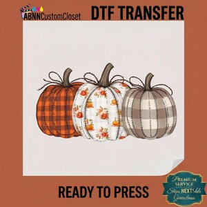 May include: A DTF transfer featuring three pumpkins with stitched details. The left pumpkin has an orange and black buffalo plaid pattern, the center one is white with orange floral accents, and the right one has a grey and white buffalo plaid pattern.